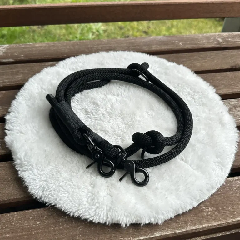 Eine Hundeleine aus schwarzem Tauseil mit schwarzer Takelung auf einem weißen, runden und flauschigen Kissen, das auf einer Holzbank liegt.