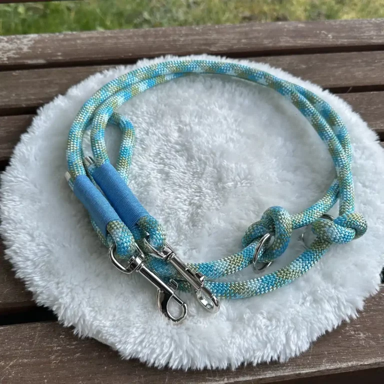 Eine Hundeleine aus blauem Tauseil mit blauer Takelung auf einem weißen, runden und flauschigen Kissen, das auf einer Holzbank liegt.