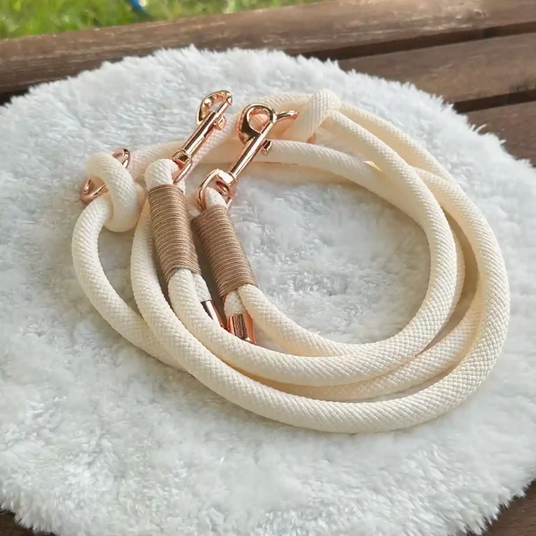 Eine Hundeleine aus beigem Tauseil mit beige-brauner Takelung auf einem weißen, runden und flauschigen Kissen, das auf einer Holzbank liegt.