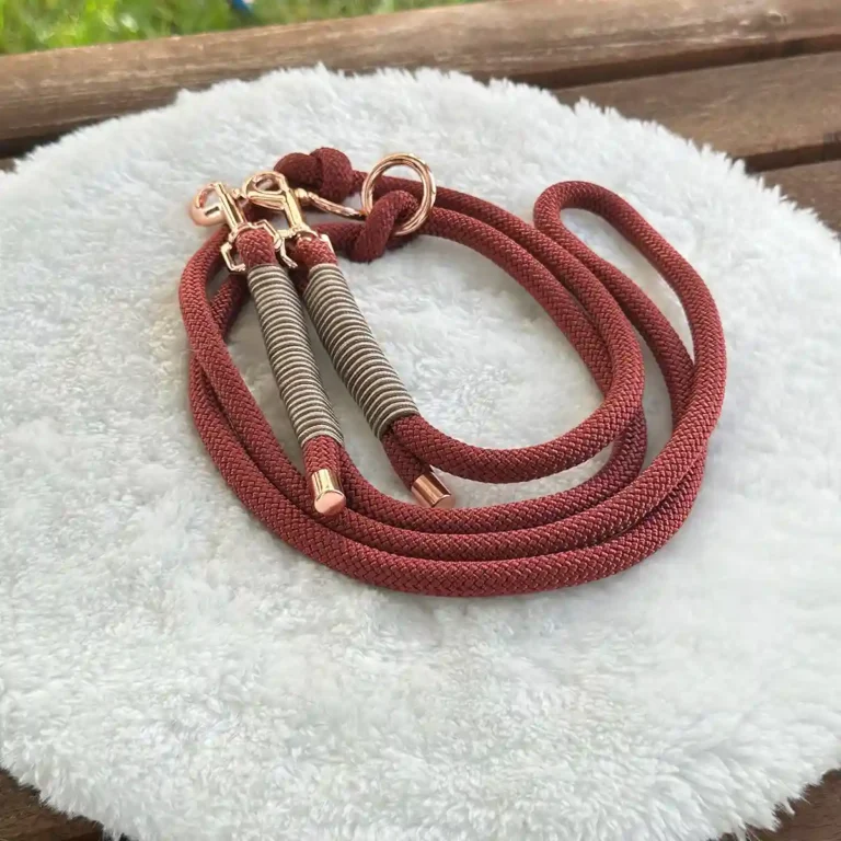 Eine Hundeleine aus braunrotem Tauseil mit beiger Takelung auf einem weißen, runden und flauschigen Kissen, das auf einer Holzbank liegt.