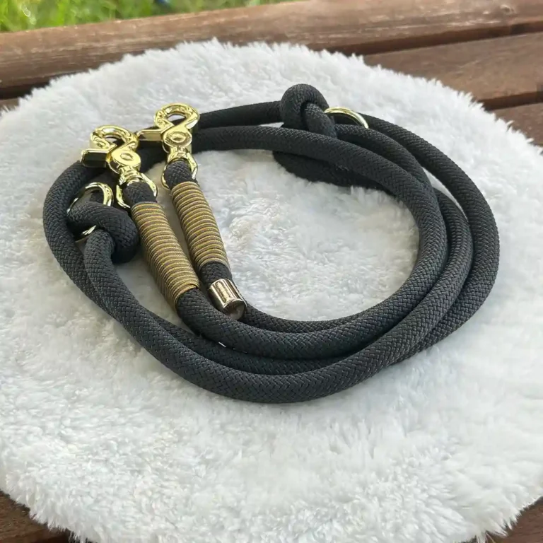 Eine Hundeleine aus dunkelgrauem Tauseil mit goldener Takelung auf einem weißen, runden und flauschigen Kissen, das auf einer Holzbank liegt.