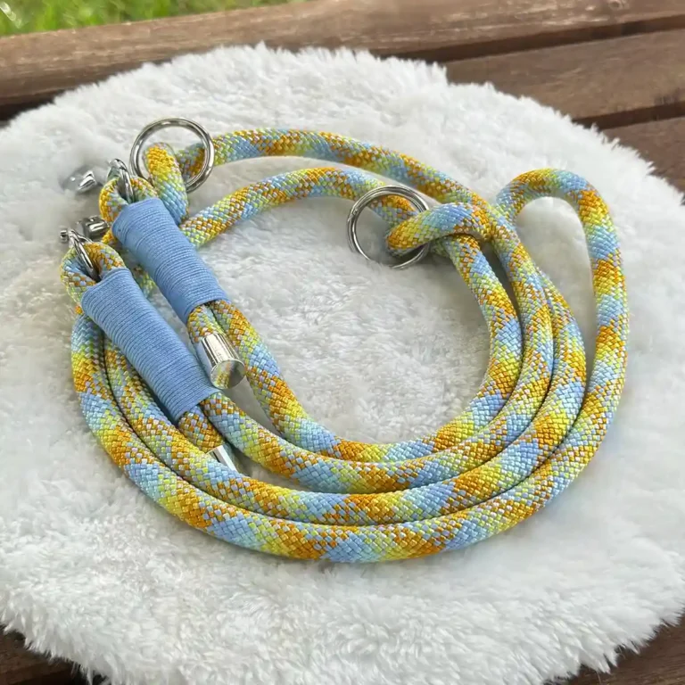 Eine Hundeleine aus gelb-hellblauem Tauseil mit hellblauer Takelung auf einem weißen, runden und flauschigen Kissen, das auf einer Holzbank liegt.
