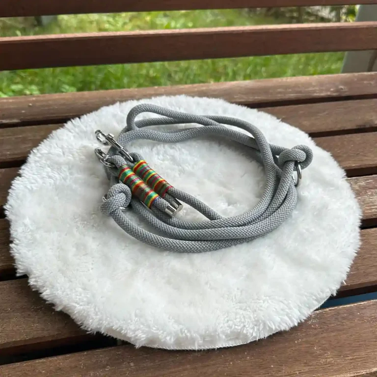 Eine Hundeleine aus grauem Tauseil mit bunter Takelung auf einem weißen, runden und flauschigen Kissen, das auf einer Holzbank liegt.