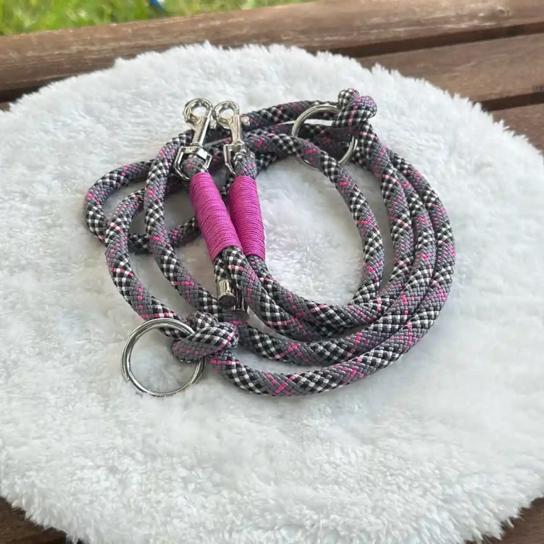 Eine Hundeleine aus grau-pinkem Tauseil mit pinker Takelung auf einem weißen, runden und flauschigen Kissen, das auf einer Holzbank liegt.