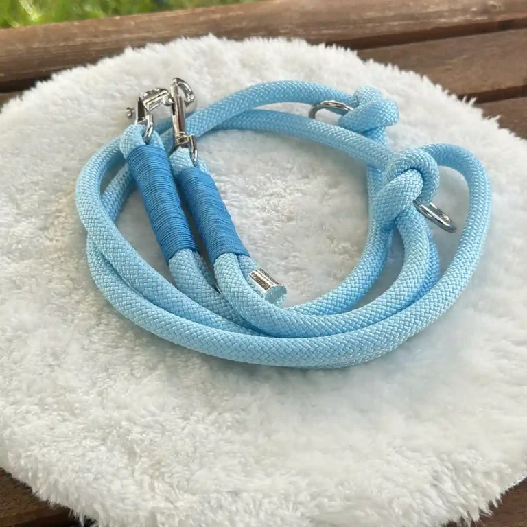 Eine Hundeleine aus hellblauem Tauseil mit blauer Takelung auf einem weißen, runden und flauschigen Kissen, das auf einer Holzbank liegt.
