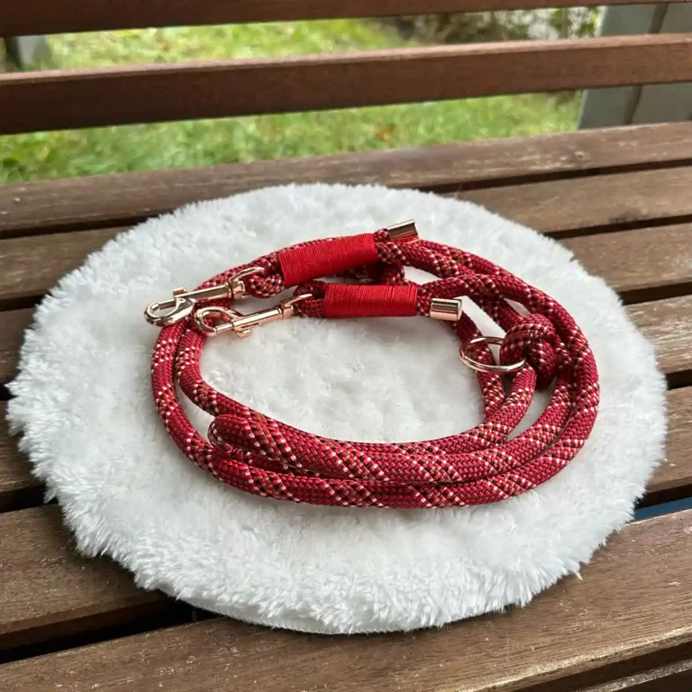 Eine Hundeleine aus rot-weißem Tauseil mit roter Takelung auf einem weißen, runden und flauschigen Kissen, das auf einer Holzbank liegt.